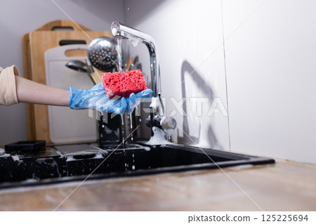 A Bright Red Sponge Being Used for Cleaning Dishes in a Modern Kitchen Sink Under Running Water with Bubbles and Kitchen Utensils in the Background 125225694
