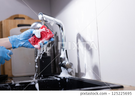 Vibrant Pink Kitchen Sponge Held in Gloved Hand Under Bubbling Water from Faucet While Cleaning Dishes in Modern Kitchen Setting 125225695