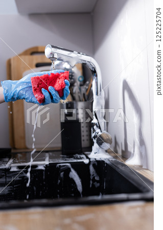 Bright Red Sponge Being Used to Clean a Sink in Modern Kitchen with Water and Soap Bubbles Flowing from the Faucet and Various Kitchen Utensils in the Background 125225704