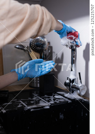 Person Cleaning Kitchen Sink with Soap and Sponge Wearing Blue Gloves, Focused on Cleaning Task in Neatly Organized Kitchen Environment 125225707