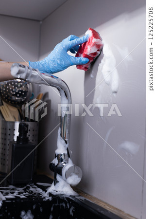 Person Wearing Blue Gloves Cleaning a Wall with a Red Sponge and Soap Foam in a Modern Kitchen Setting with a Shiny Faucet and Kitchen Utensils in Background Person Wearing Blue Gloves Cleaning a Wall with a Red Sponge and Soap Foam in a Modern Kitchen Setting with a Shiny Faucet and Kitchen Utensils in Background 125225708