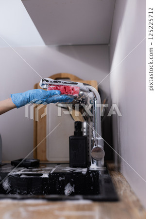 Cleaning Tools in Action: A Gloved Hand Holding a Pink and White Scrubbing Sponge Over a Soapy Kitchen Sink with a Modern Aesthetic and Various Kitchen Utensils Nearby 125225712