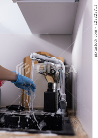 A close-up view of a hand using a faucet with soapy water, emphasizing cleanliness while preparing to do the dishes in a cozy kitchen setting with utensils in the background. 125225713