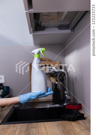 A Clean Kitchen Scene Featuring a Hand Holding a Spray Bottle for Household Cleaning and Sanitizing Tasks in a Modern Kitchen Environment with Essential Utensils 125225733