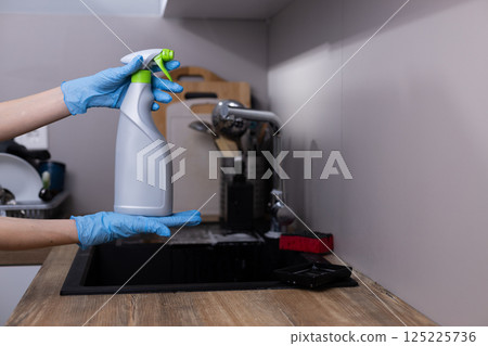 Hands in Blue Gloves Holding a Cleaning Spray Bottle in a Modern Kitchen Setting Focused on Hygiene and Cleanliness with Kitchen Utensils Visible in the Background. 125225736