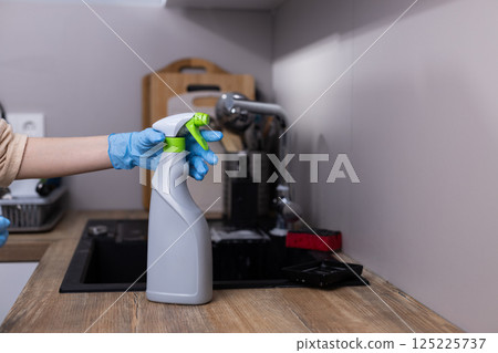 Hand in Blue Gloves Holding a Spray Bottle for Cleaning on a Wooden Countertop in a Modern Kitchen with a Sink and Kitchen Utensils in the Background 125225737