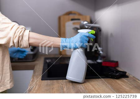 A Person in Gloves Preparing to Clean a Kitchen Surface with a Spray Bottle, Showcasing a Household Cleaning Routine with Attention to Hygiene and Cleanliness in a Modern Home Environment A Person in Gloves Preparing to Clean a Kitchen Surface with a Spray Bottle, Showcasing a Household Cleaning Routine with Attention to Hygiene and Cleanliness in a Modern Home Environment 125225739