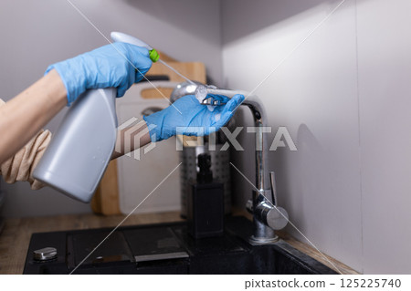 Engaging in the Act of Cleaning: A Close-Up of a Hand with Blue Gloves Spraying Cleaning Solution on a Kitchen Faucet with Utensils in the Background Engaging in the Act of Cleaning: A Close-Up of a Hand with Blue Gloves Spraying Cleaning Solution on a Kitchen Faucet with Utensils in the Background 125225740
