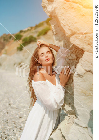A woman in a white dress is standing on a rocky beach. Concept of serenity and tranquility, as the woman is enjoying the natural beauty of the beach. 125225840