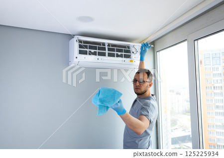 Man Performing Maintenance and Cleaning on Air Conditioning Unit in Modern Apartment, Ensuring Optimal Functionality and Fresh Air Quality in Urban Environment Man Performing Maintenance and Cleaning on Air Conditioning Unit in Modern Apartment, Ensuring Optimal Functionality and Fresh Air Quality in Urban Environment 125225934