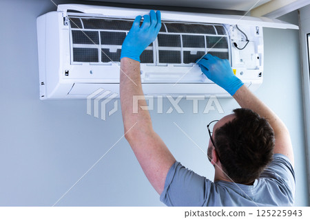 Man in Grey Shirt and Blue Gloves Performing Maintenance on Air Conditioning Unit in Modern Interior Environment for Optimal Cooling Efficiency and Clean Air Quality Improvement 125225943