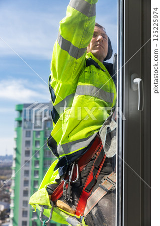 A Dedicated Window Cleaner in a Yellow Safety Suit Working on a High-Rise Building While Harnessed for Safety and Using Professional Equipment to Ensure a Flawless Shine 125225974