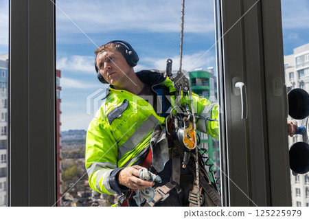 Skilled Window Cleaner Working on High-Rise Building, Captured in Bright Daylight, Wearing Safety Gear and Headphones for Enhanced Focus and Protection 125225979
