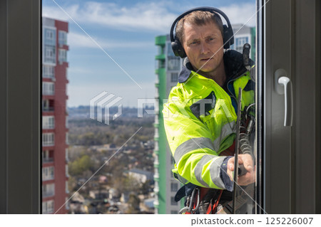 A Professional Window Cleaner Safely Working on High-Rise Building, Demonstrating Expertise in Urban Maintenance and Safety Protocols While Wearing Protective Gear 125226007