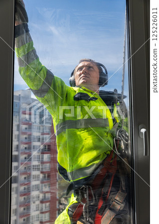 Professional Window Cleaner at Work, Wearing Safety Gear and Focused on Cleaning Glass, Showcasing Skill and Care in High Rise Building Maintenance 125226011