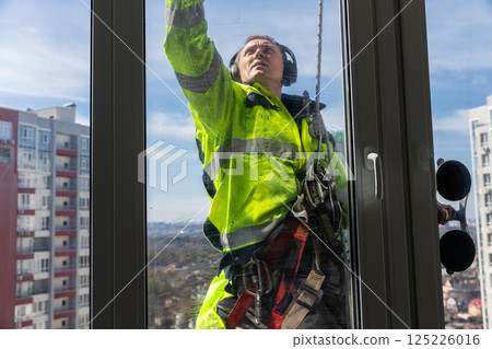 Professional Window Cleaner in Bright Safety Gear Cleaning a High-Rise Building with a Scenic Urban Background Under a Clear Blue Sky on a Sunny Day 125226016