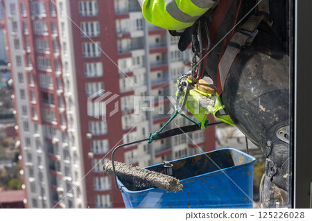 Dynamic Construction Scene: Worker in Safety Gear Painting a High-Rise Building with City View in the Background, Showcasing the Intersection of Urban Life and Skilled Labor Dynamic Construction Scene: Worker in Safety Gear Painting a High-Rise Building with City View in the Background, Showcasing the Intersection of Urban Life and Skilled Labor 125226028