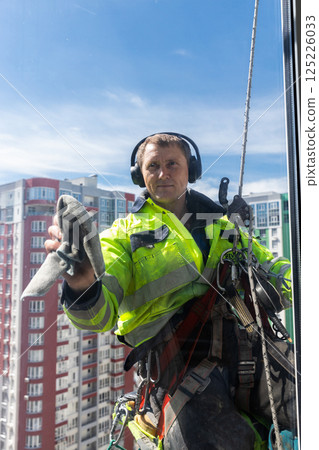 Professional Window Cleaner at Work: A Skilled Worker in High-Rise Building Wearing Safety Gear and Headphones While Cleaning Windows in a Bright Blue Sky 125226033