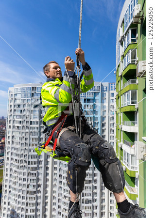A Dedicated Worker Performs High-Rise Maintenance While Suspended in the Air, Secured by Ropes Near Tall Modern Apartment Buildings on a Clear Sunny Day 125226050