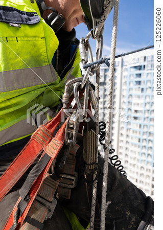 A close-up of a construction worker's safety equipment as they prepare for a climbing ascent, showcasing harnesses, carabiners, and a clear blue sky backdrop for safety training purposes. 125226060