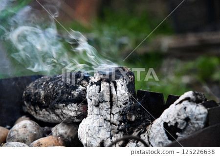 Flames and smoke rise from burning wood in a rustic outdoor fire pit as embers glow under the charred 125226638