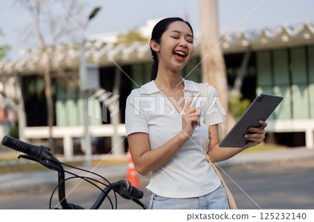 Sustainable Transportation. Young woman enjoying her bicycle ride while using a tablet in a park. 125232140