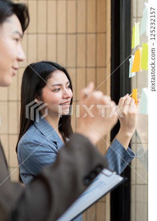 Team members collaborating on project ideas with sticky notes on a glass board. 125232157