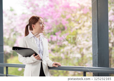 Reflective businesswoman holding files and gazing at blooming flowers in a modern office setting 125232165