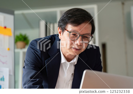 Elderly professional engaged in online work with a laptop and financial data. Elderly professional engaged in online work with a laptop and financial data. 125232338