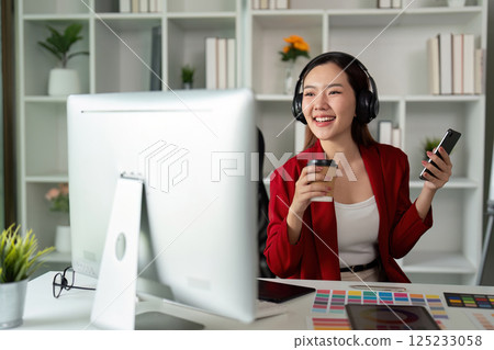 Joyful Design and Relaxation. A smiling graphic designer enjoys a coffee while creatively engaging with her digital tools in a vibrant workspace. 125233058