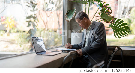 Focused Work and Reflection. A senior man writing notes while observing a serene outdoor view from a modern office. 125233142