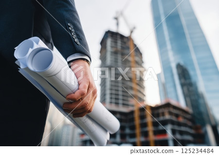 Architect in Suit Holding Rolled Blueprints at Construction Site in City Setting 125234484