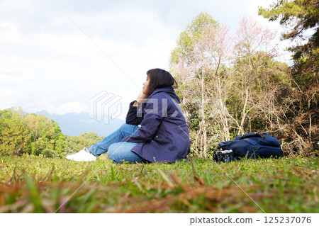 Traveler Asian young girl with bag and camera happy and sitting on green lawn field on the hill with valley Doi Luang Chiang Dao mountain view, Chiang Mai Province of Thailand 125237076