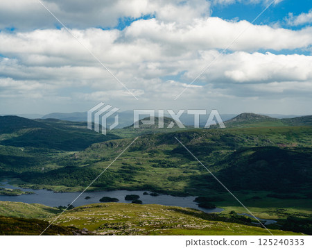 High-angle shot showing diverse terrain. Green hills, dark forests, and a body of water. The sky is mostly cloudy. 125240333