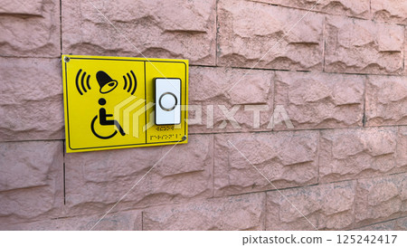 Bright yellow accessibility sign with ringing bell symbol and wheelchair icon, mounted on pink stone wall next to white button with braille for calling assistance at accessible entrance Bright yellow accessibility sign with ringing bell symbol and wheelchair icon, mounted on pink stone wall next to white button with braille for calling assistance at accessible entrance 125242417