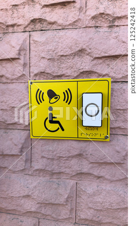 Vertical photo of bright yellow accessibility call button with tactile Braille and wheelchair icon mounted on textured pink wall, emphasizing inclusive access and support for disabled persons Vertical photo of bright yellow accessibility call button with tactile Braille and wheelchair icon mounted on textured pink wall, emphasizing inclusive access and support for disabled persons 125242418