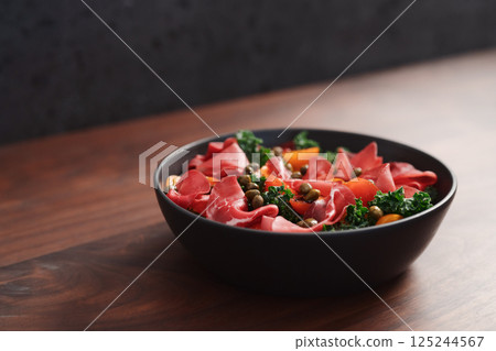 Salad with kale, bresaola and cherry tomatoes decorated with capers in black bowl on walnut wood table 125244567