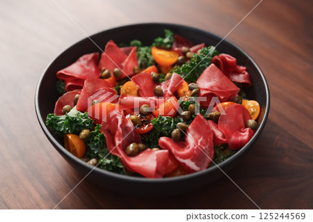 Salad with kale, bresaola and cherry tomatoes decorated with capers in black bowl on walnut wood table Salad with kale, bresaola and cherry tomatoes decorated with capers in black bowl on walnut wood table 125244569