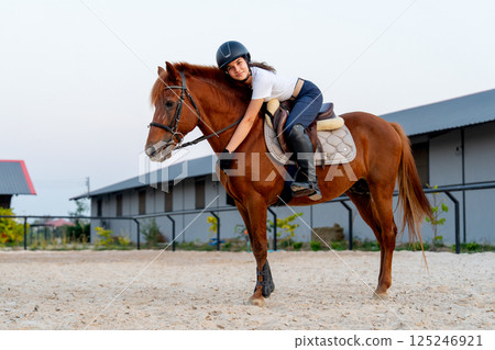 Side view of teen girl stay on the horse and bend down to make horse to relax before train or practice in the arena. 125246921