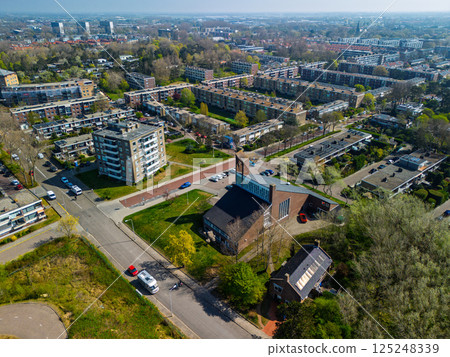 Aerial view of a residential neighborhood with a church at the center, surrounded by apartment buildings, green spaces, roads, and trees under a clear sky. 125248339