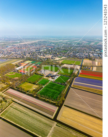 Aerial footage shows colorful tulip fields, greenhouses, and canals stretching across the flat Dutch landscape, with a lake and distant towns visible under an overcast sky. 125248343