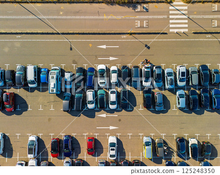 Aerial view of a large, organized parking lot filled with colorful cars. Bright sunlight casts long shadows, highlighting the neat rows and road markings surrounding the area. 125248350