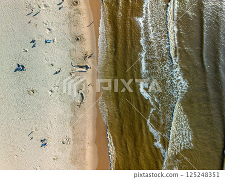Aerial view of a beach where gentle waves meet the sandy shore. People are scattered across the sand, some building sandcastles, others relaxing and enjoying the sunny day. 125248351