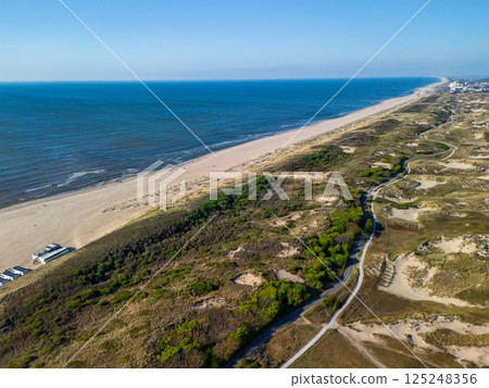 A stunning aerial view of a long, sandy beach stretching along the coast, bordered by dunes and greenery, with a clear blue sky and calm sea creating a peaceful, natural atmosphere. 125248356