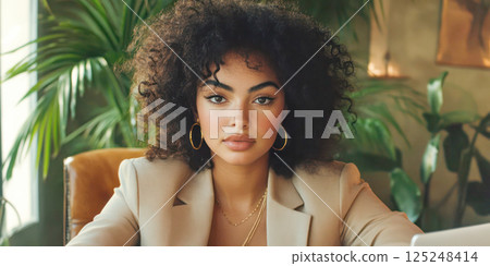 Young woman with curly hair and gold hoop earrings in a beige blazer working in a stylish office. 125248414