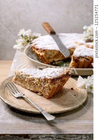 Homemade pie Pastiera Napolitano slice with powdered sugar on ceramic plate. Serving knife fork white flowers. Rustic wooden background. Delicious dessert. Homemade pie Pastiera Napolitano slice with powdered sugar on ceramic plate. Serving knife fork white flowers. Rustic wooden background. Delicious dessert. 125254771
