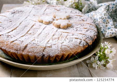 Delicious homemade pie Pastiera Napolitano with powdered sugar dusting and flowershaped decorations on top. Served on ceramic plate with white spring blossoms around rustic wooden table. 125254774