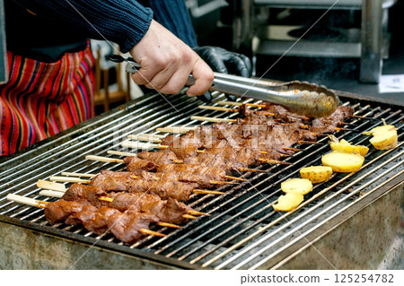 Chef grilling meat skewers and potatoes on barbecue. Hand holds tongs preparing traditional kebabs on street food festival. Outdoor cooking process. Chef grilling meat skewers and potatoes on barbecue. Hand holds tongs preparing traditional kebabs on street food festival. Outdoor cooking process. 125254782
