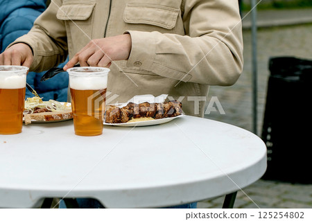 Person enjoying grilled meat skewers and draft beer at outdoor table. Casual dining scene with person in beige jacket eating street food. Person enjoying grilled meat skewers and draft beer at outdoor table. Casual dining scene with person in beige jacket eating street food. 125254802