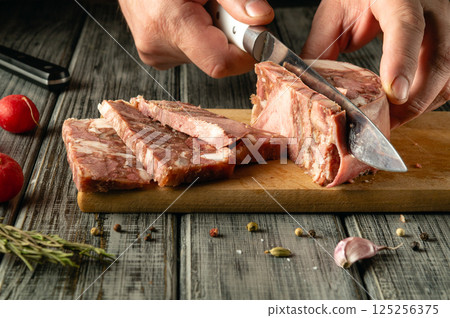 A chef skillfully slices a piece of cured meat on a wooden cutting board. Fresh herbs, spices, and garlic are scattered around, enhancing the rustic kitchen vibe during the preparation 125256375